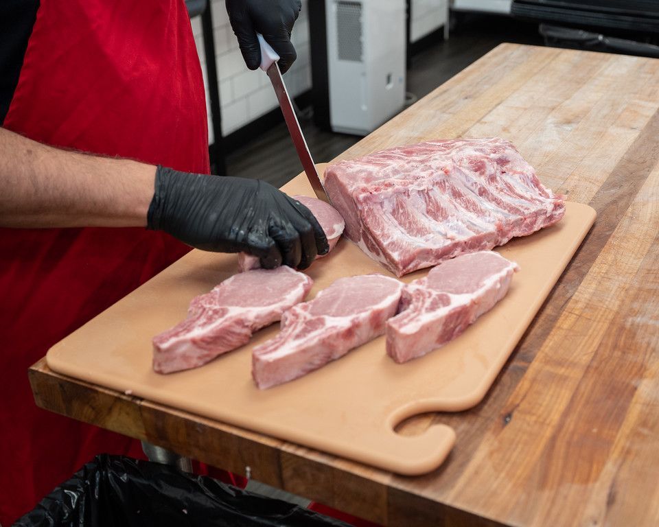 Butcher cutting pork chops on a cutting board; red apron, black gloves.