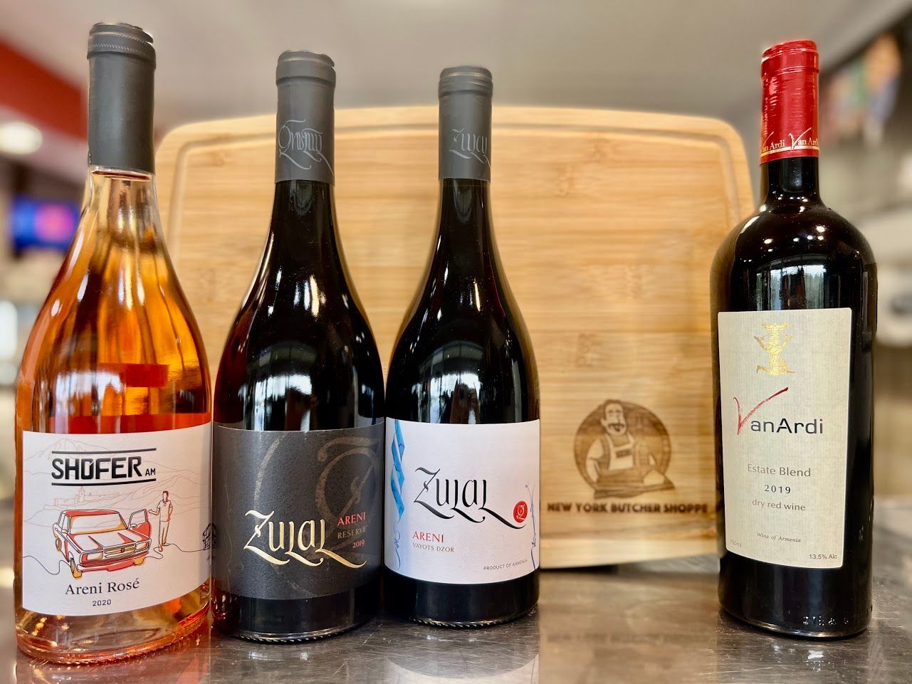 Four wine bottles sit in front of a wooden cutting board. Colors include red, pink, and black.