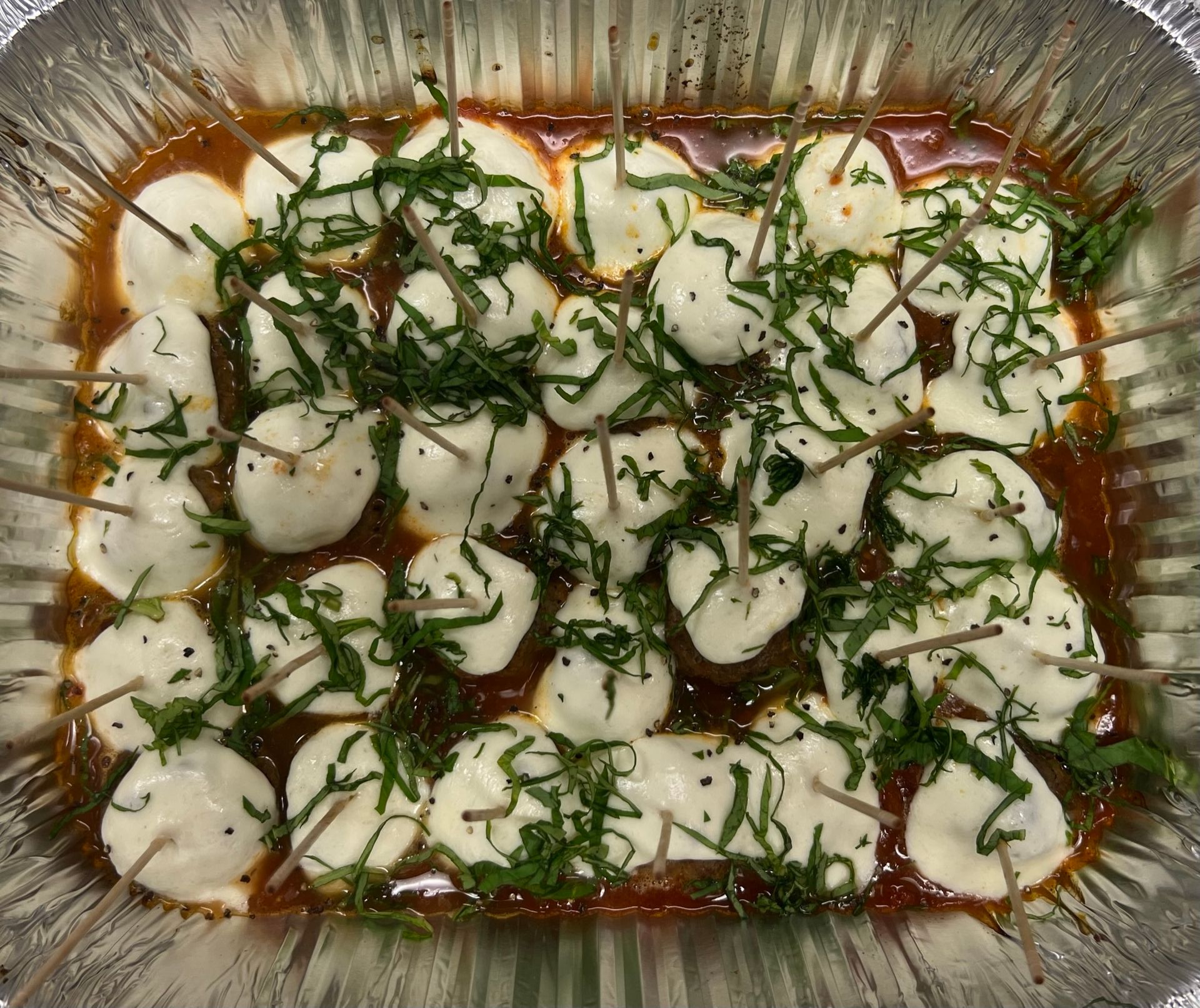 Skewered mozzarella balls in sauce, topped with green herbs, in a foil tray.