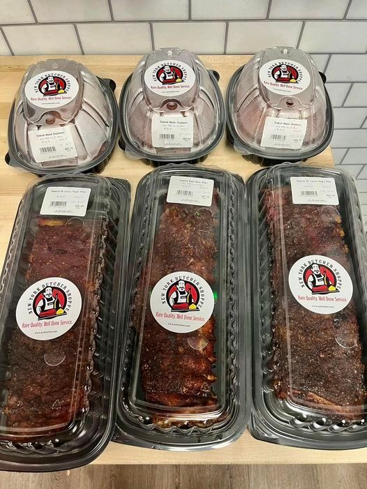 Pre-packaged meats with labels and logos sit on a wooden surface. Three rotisserie chickens sit above three rib slabs.