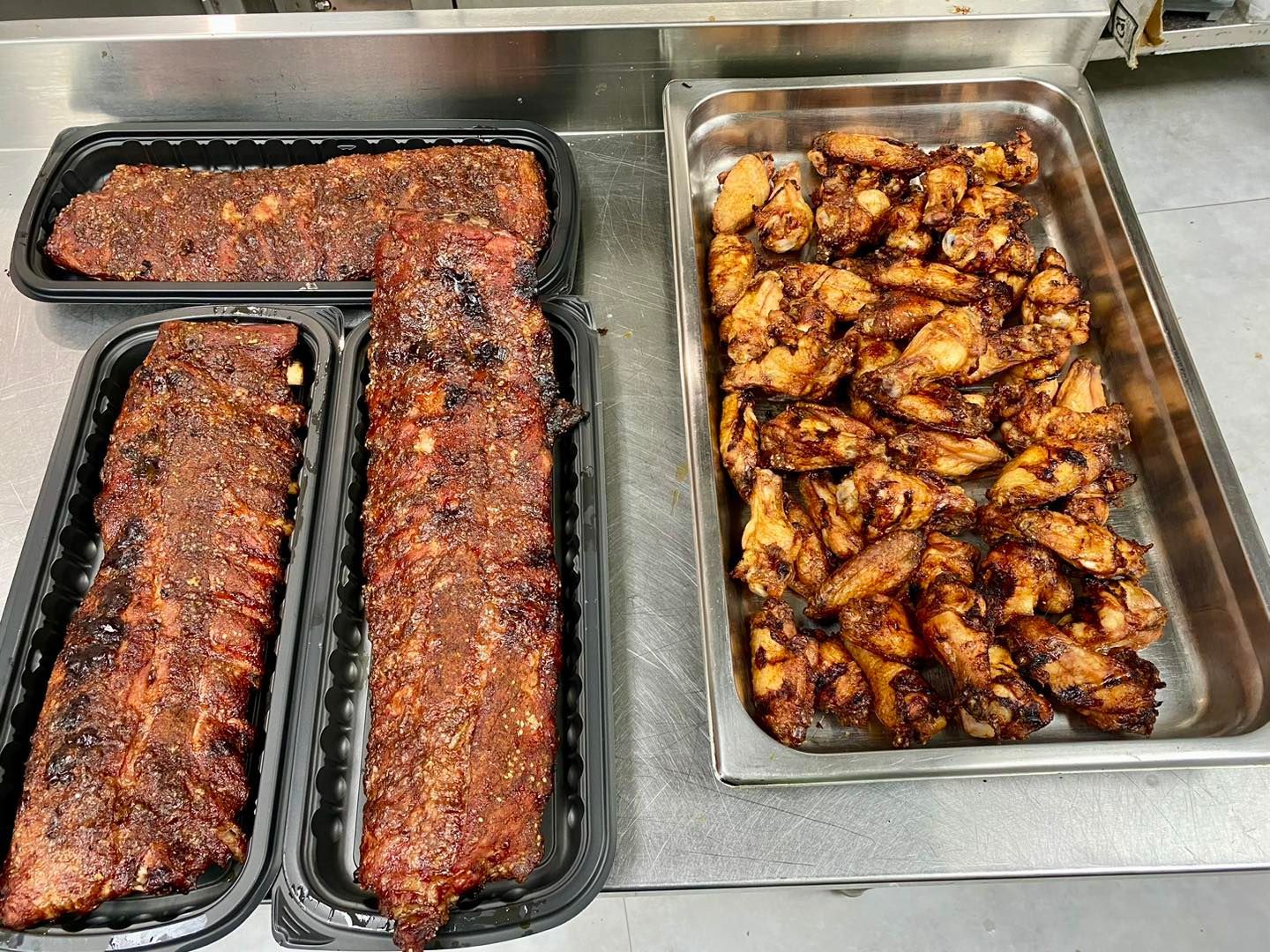 Ribs and wings in serving trays on a stainless steel surface.