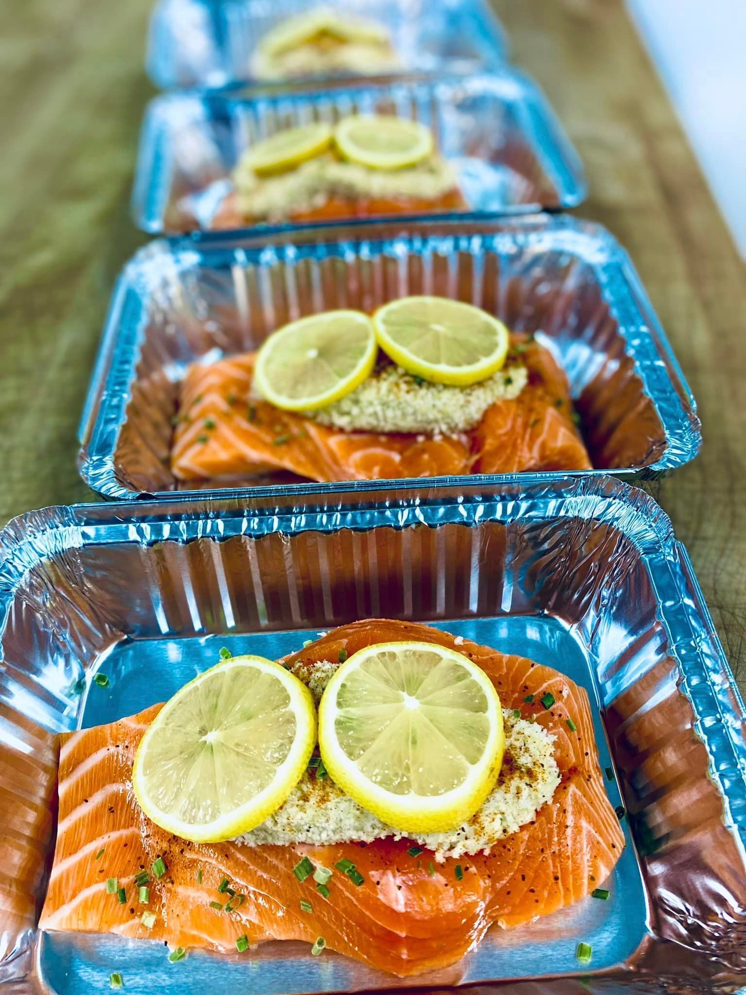 Salmon fillets in foil containers, topped with lemon slices and breadcrumbs, ready for baking.