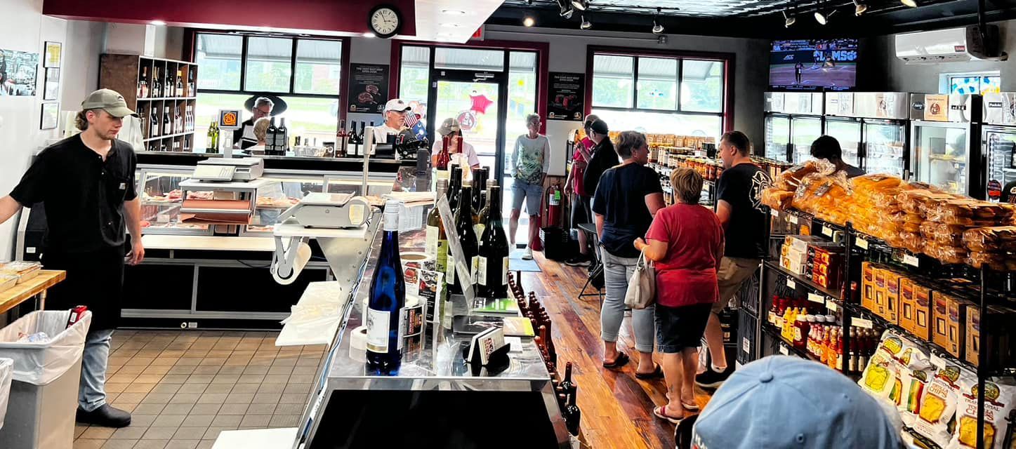A deli counter with customers and staff. A person in a hat stands by the counter; others queue to order.