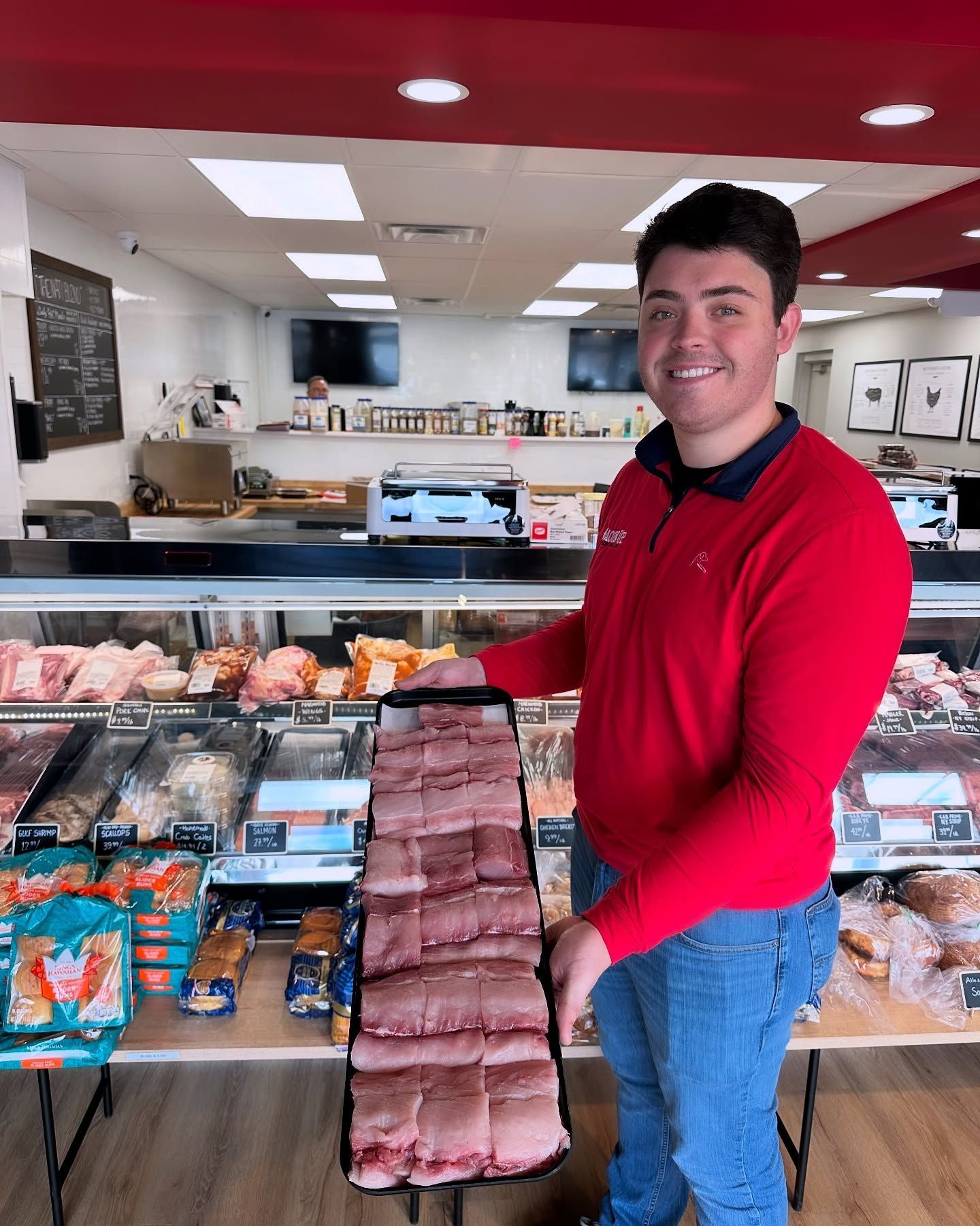 Man in red shirt holding meat in a butcher shop. He is smiling.