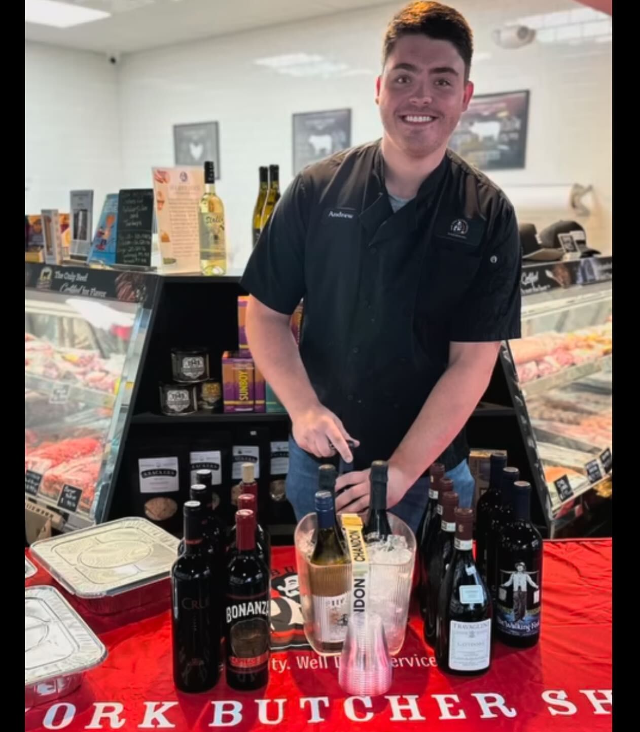 Man in butcher shop, smiling, with wine bottles on display.
