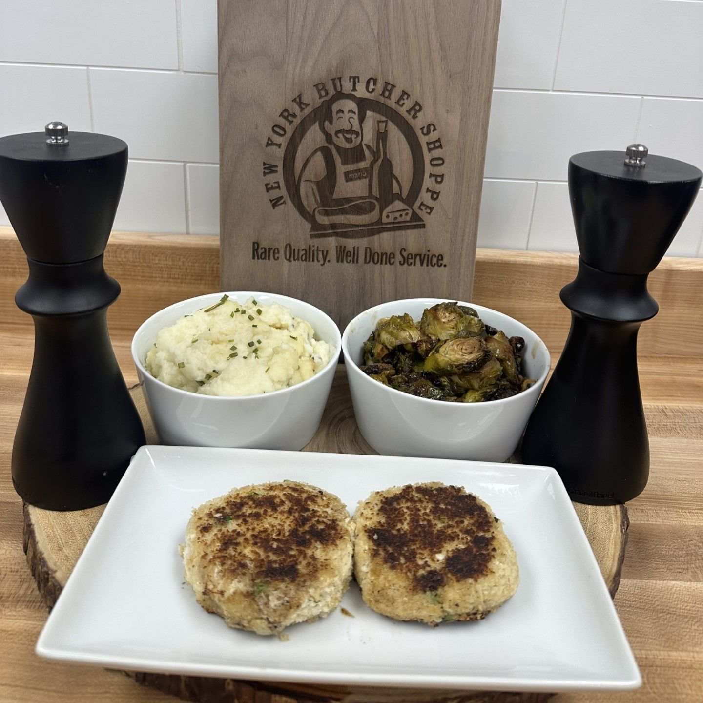 Two crab cakes, mashed potatoes, Brussels sprouts, and pepper mills, set on a wooden surface.