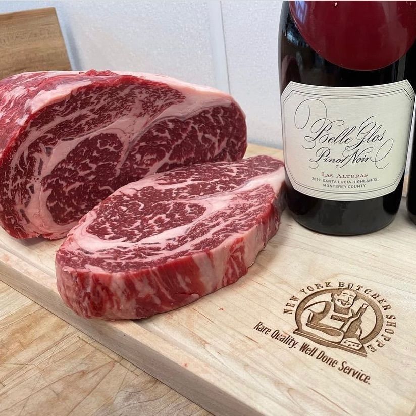 A marbled ribeye steak and a cut steak sit on a wooden cutting board with wine bottles.