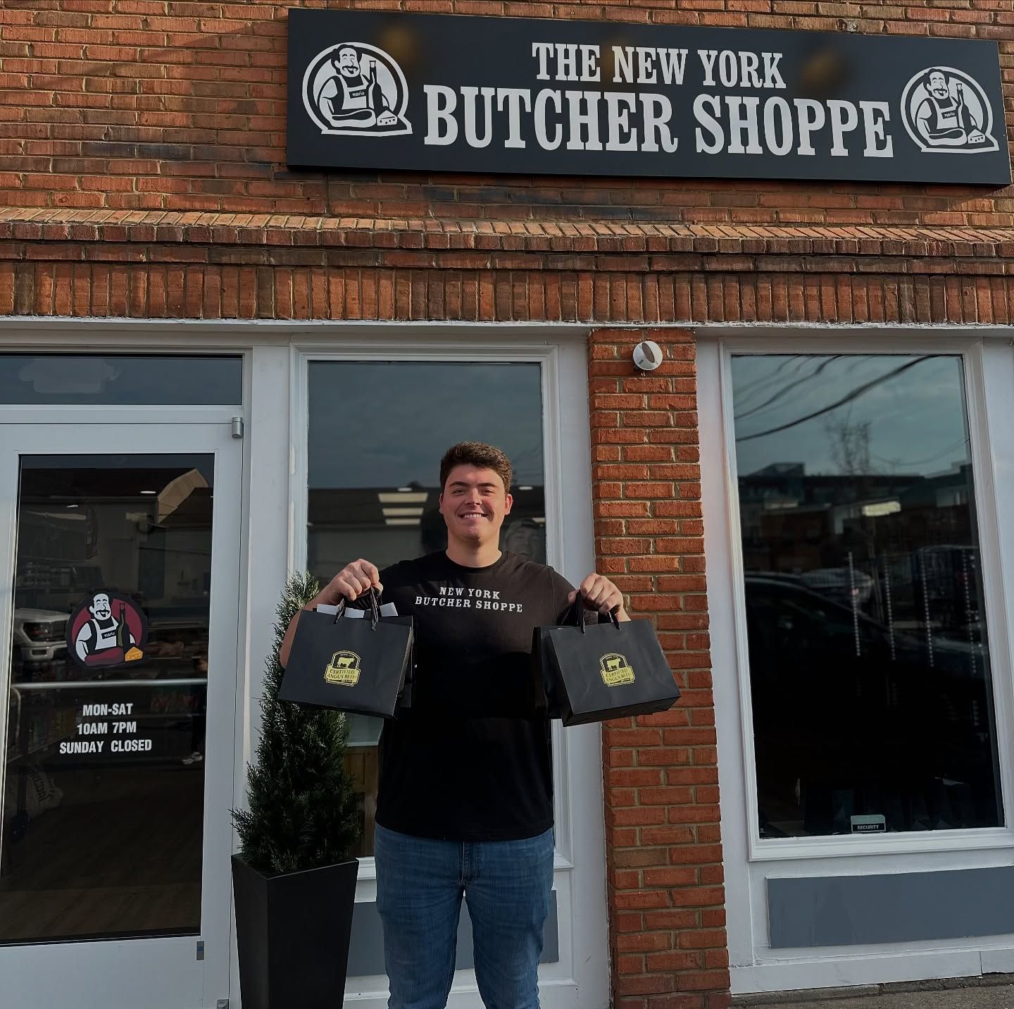 Man stands in front of the New York Butcher Shoppe holding shopping bags; brick building.