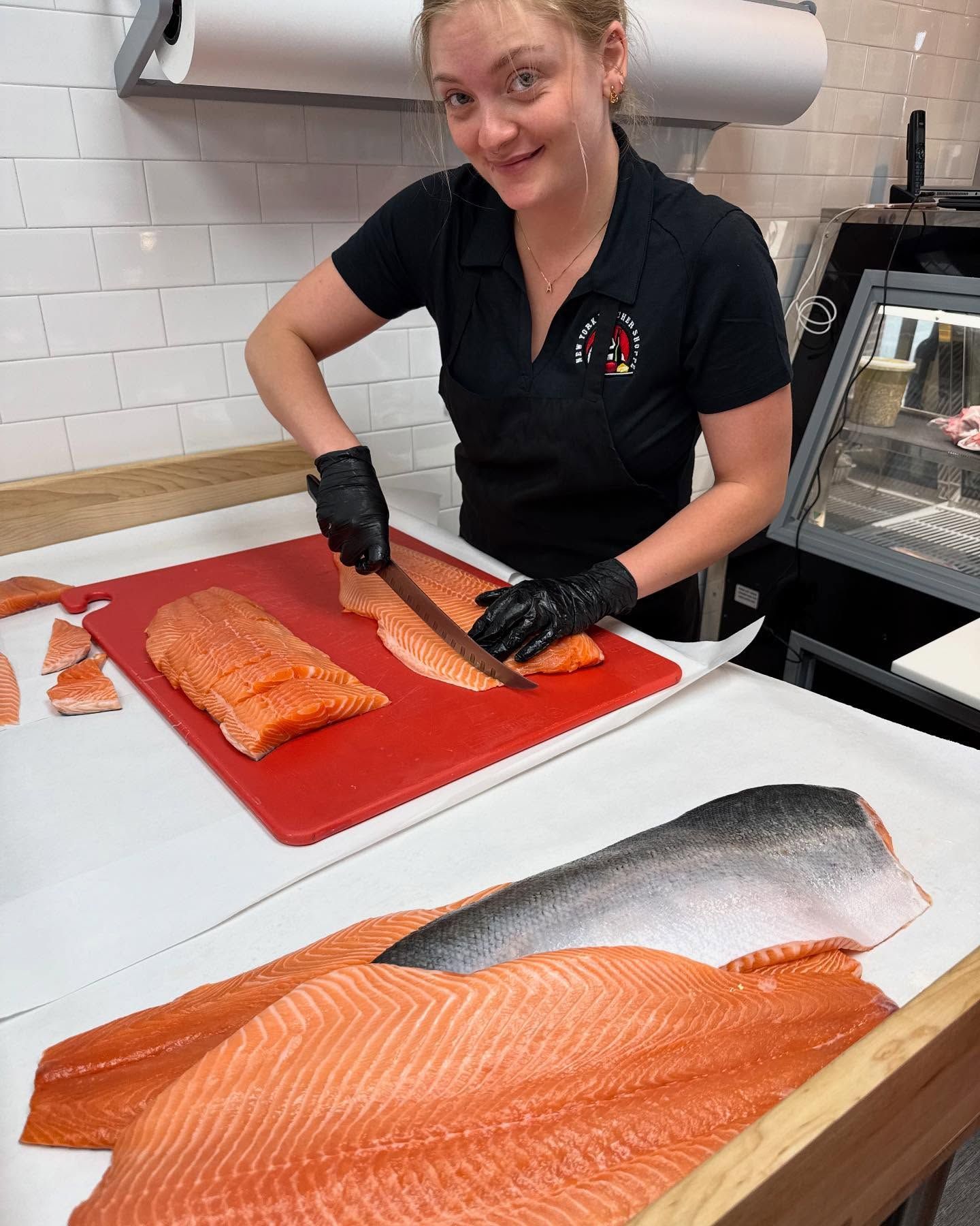 Woman slicing salmon filets on a red cutting board; she smiles, wearing a black shirt and gloves.
