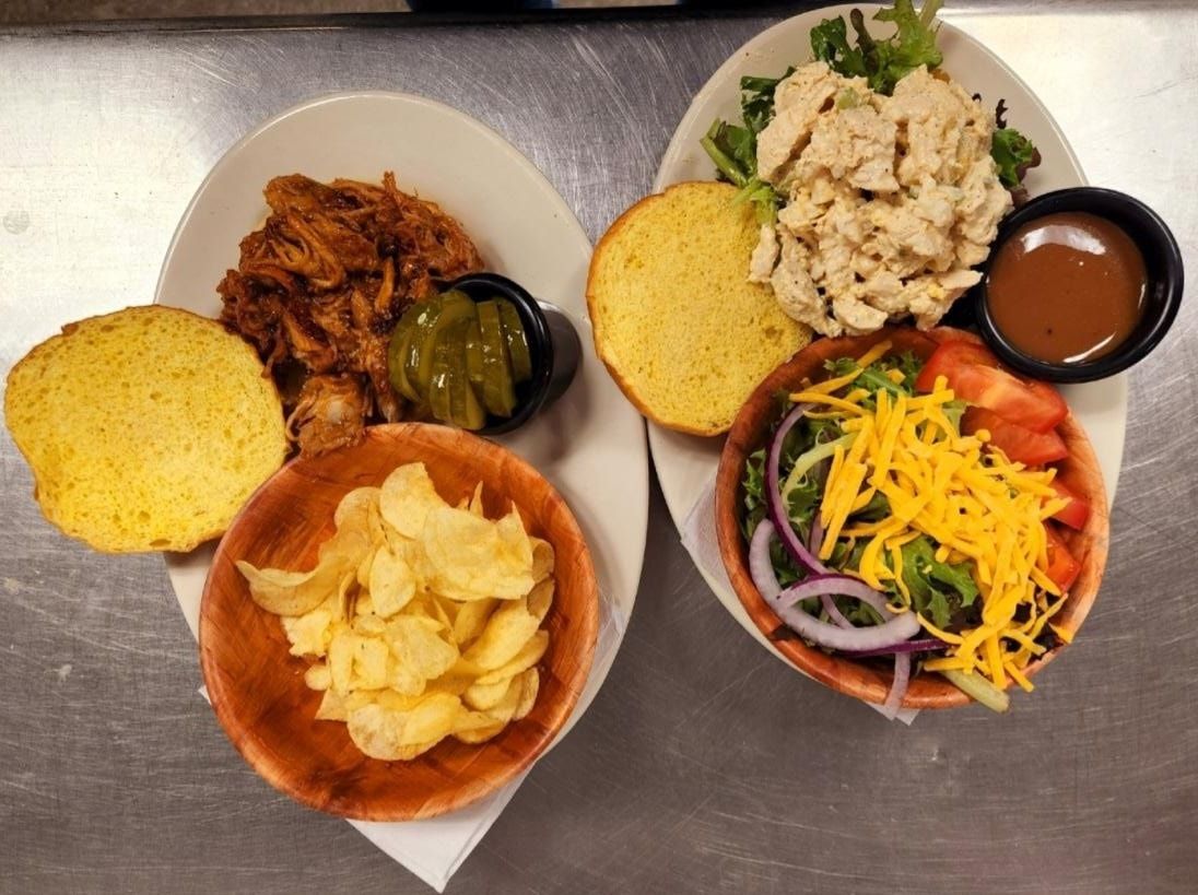 Two plates of food: pulled pork sandwich with chips and chicken salad sandwich with side salad.