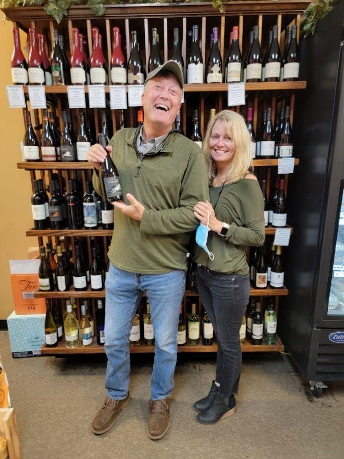 Man holding wine, woman smiling in a shop filled with wine bottles. They are smiling.