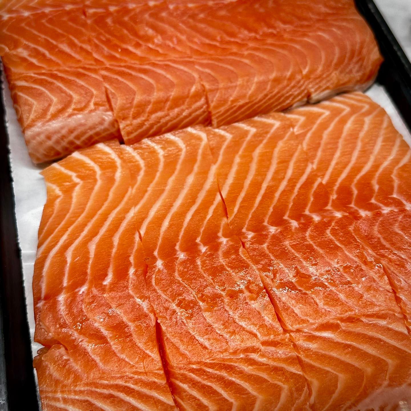 Fresh, raw salmon fillets, orange-pink with white fat lines, cut into portions, in a black tray.