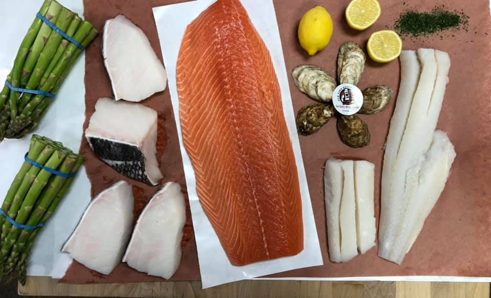 Assortment of fresh seafood: salmon, halibut, oysters, cod, asparagus, lemons, and herbs on butcher paper.