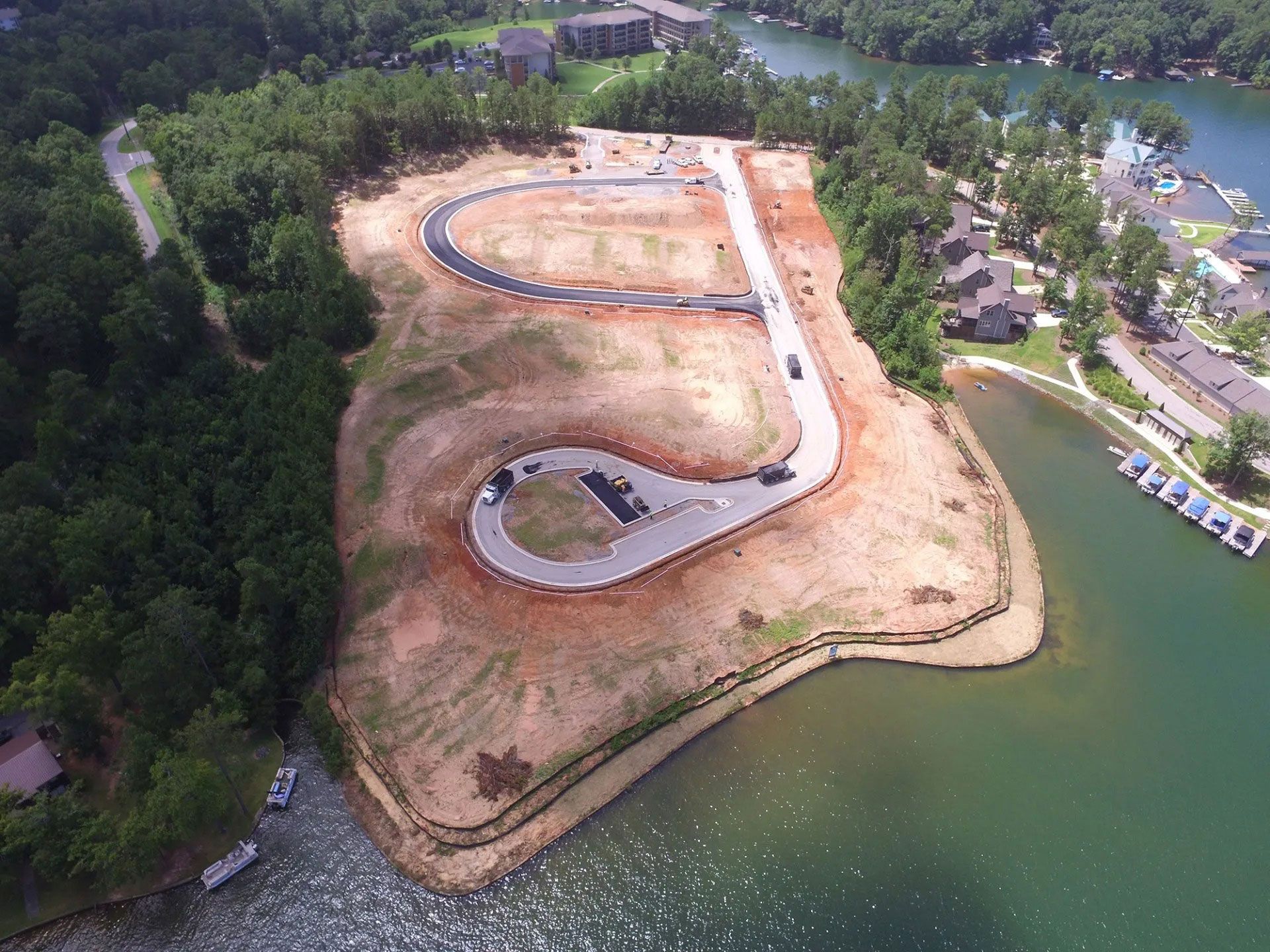Aerial view of a curved construction site on a wooded lakeshore beside green water and docks
