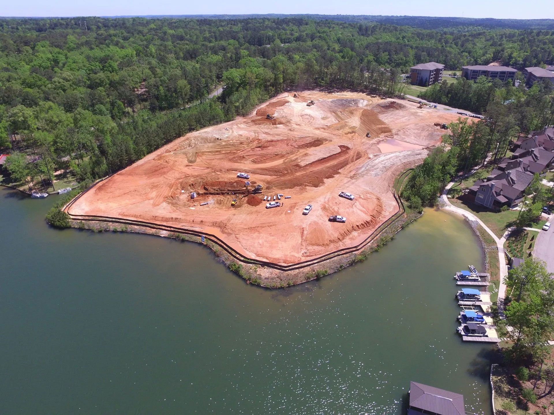 Aerial view of a cleared sandy shoreline beside green water and trees, with construction equipment onsite