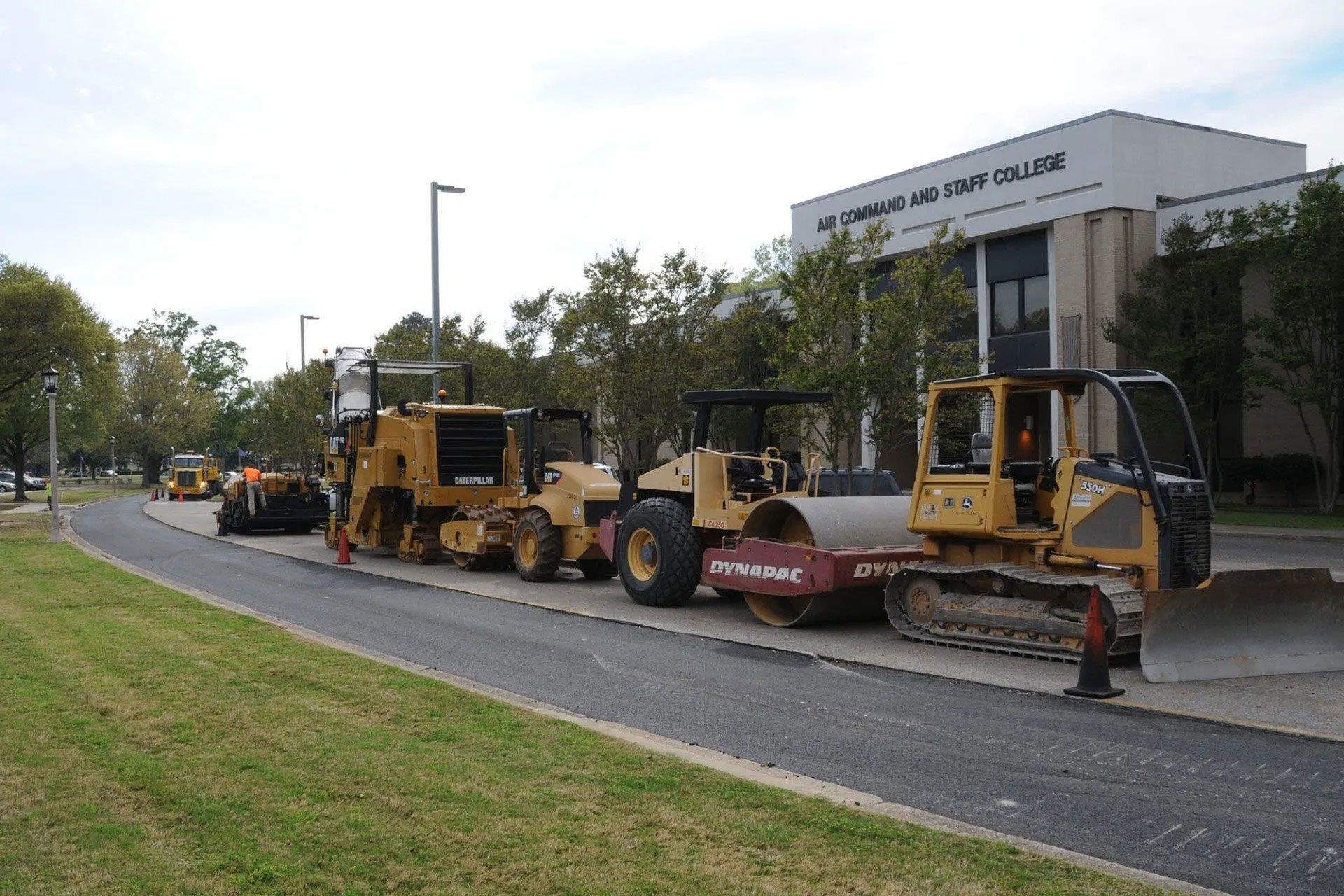 Road construction vehicles lined up beside a building on a tree-lined street