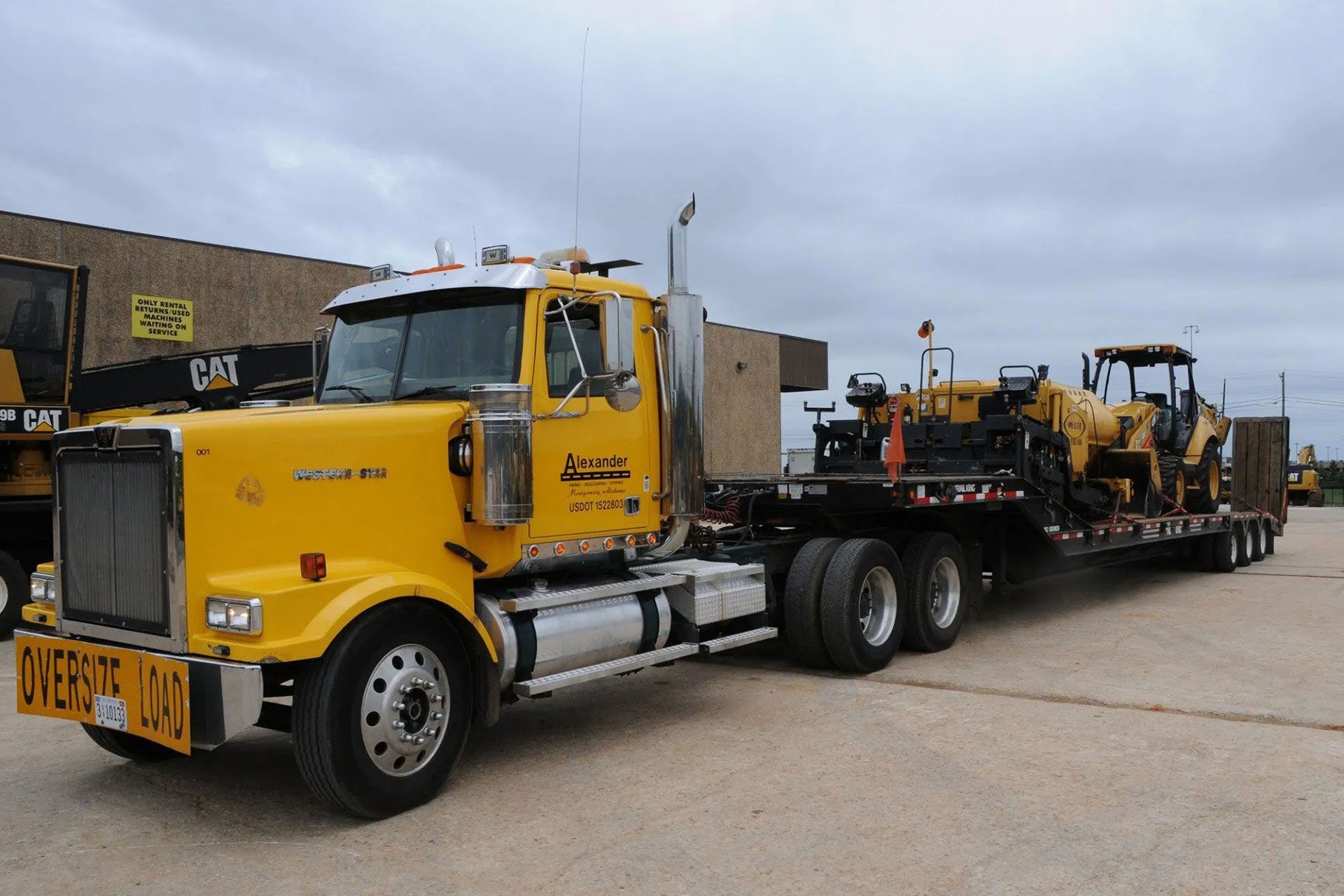 Yellow semi truck hauling heavy machinery on a flatbed trailer in an industrial lot.