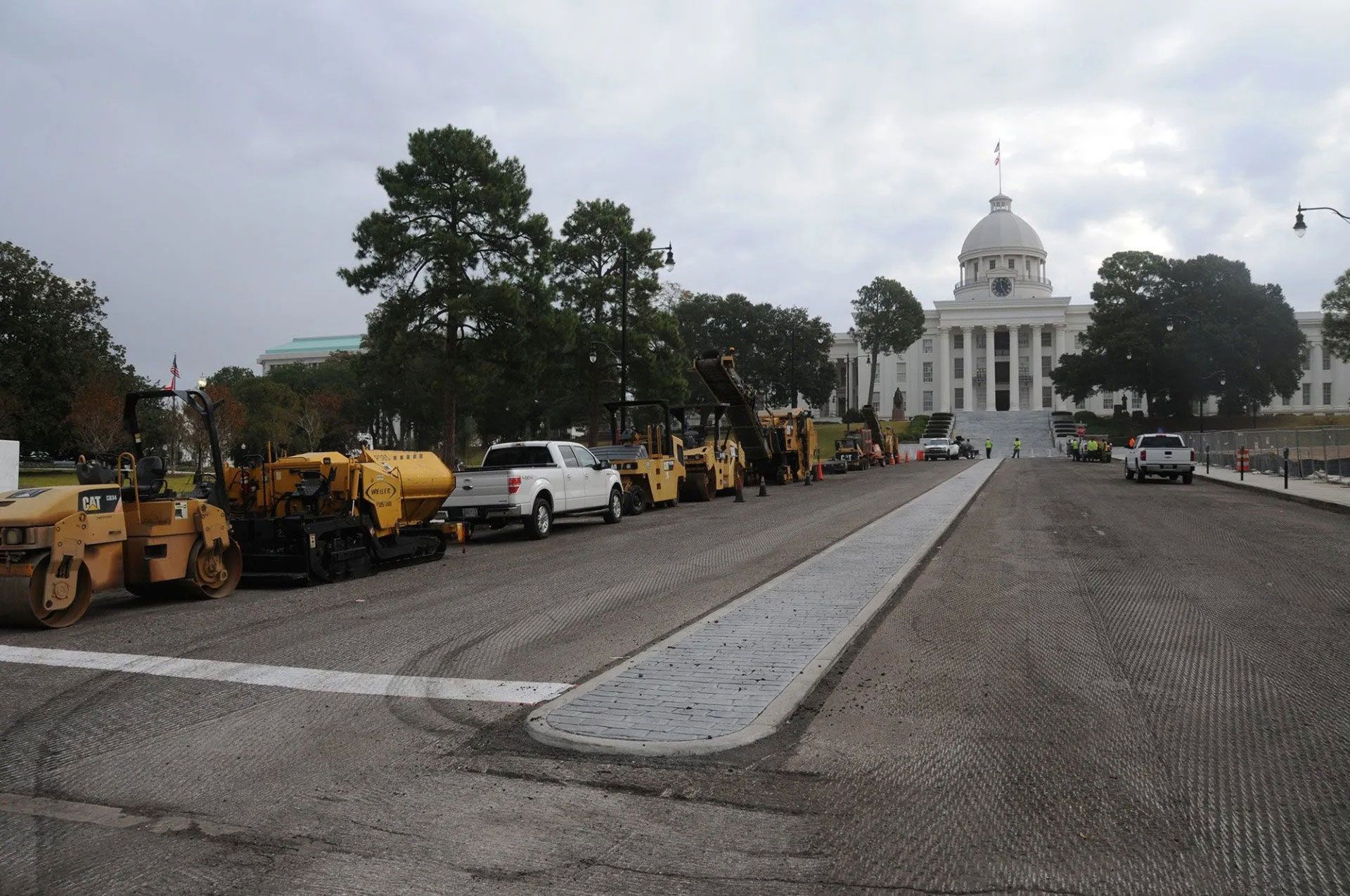 Construction vehicles lined along a road leading to a white domed capitol building in the distance.