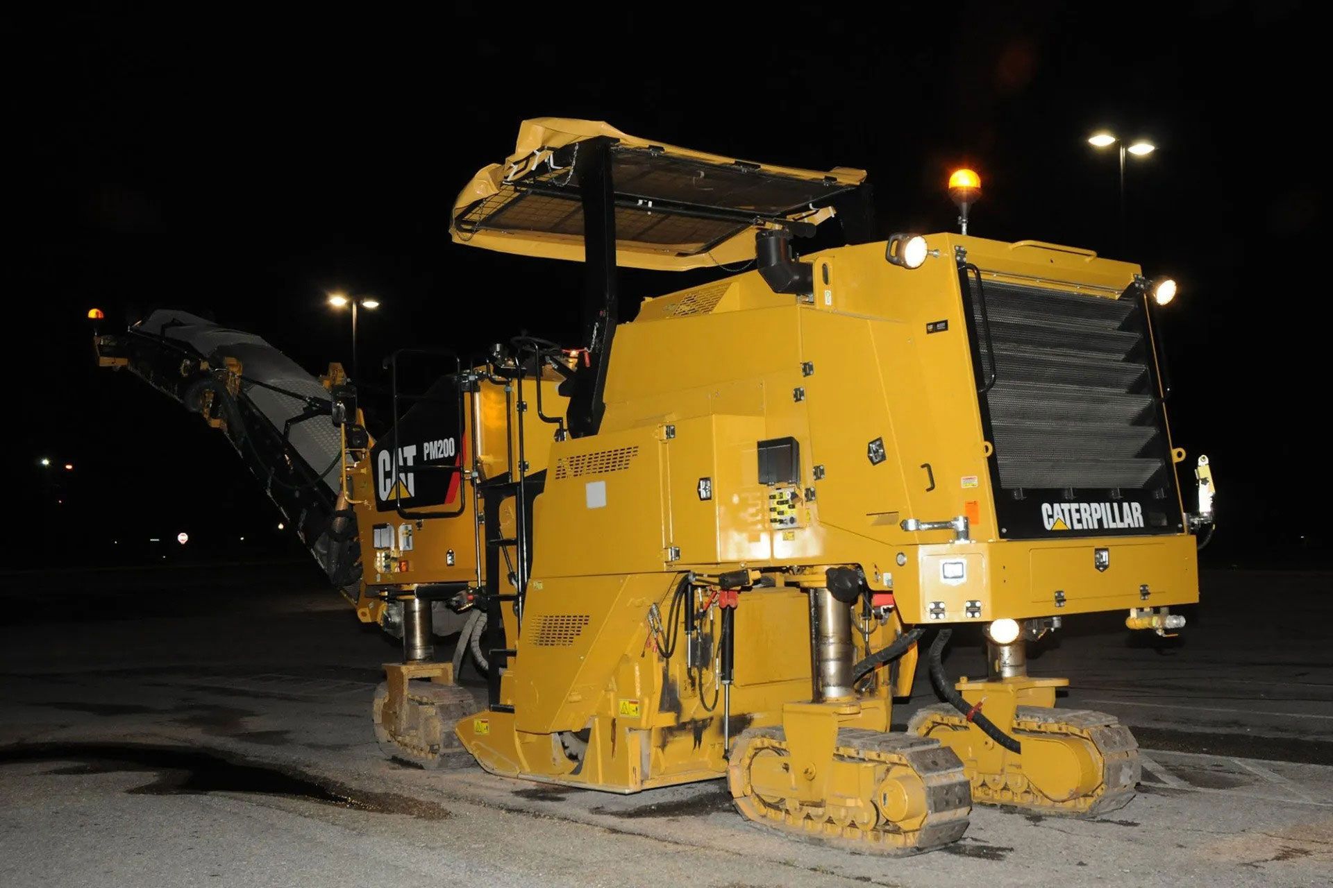 Yellow road milling machine on a dark street at night, with conveyor and cutting drum visible.