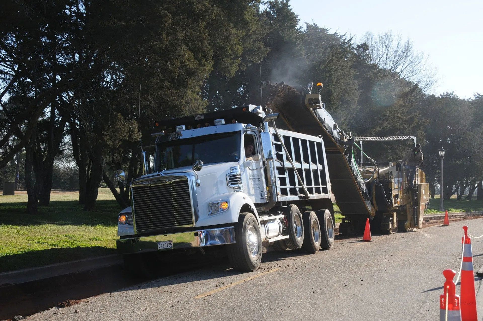 Truck with pavement equipment on a road, with orange cones and smoke in a tree-lined work zone