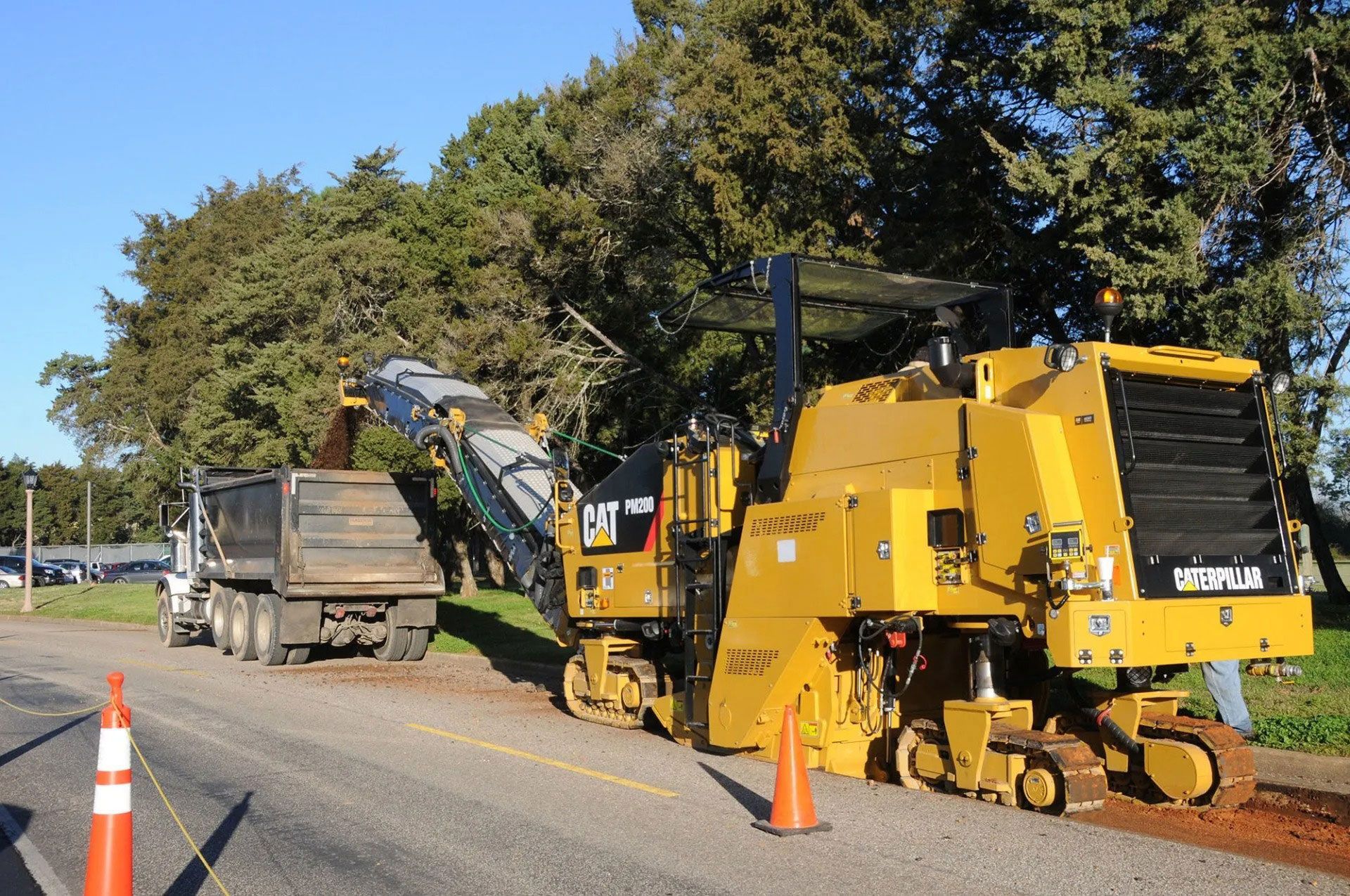 Yellow road milling machine loading asphalt into a dump truck on a roadside, with traffic cones nearby