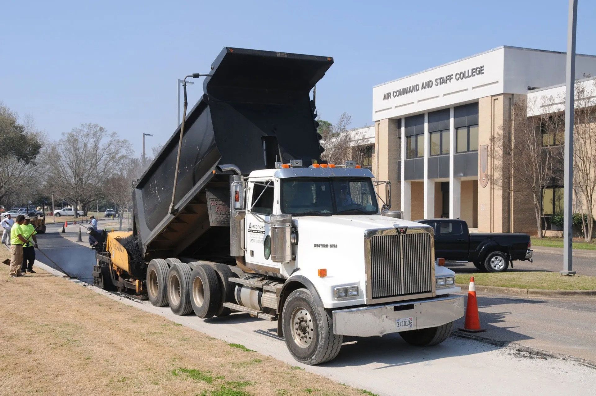 Dump truck unloading gravel beside a building, with workers and traffic cones on a sunny roadside.