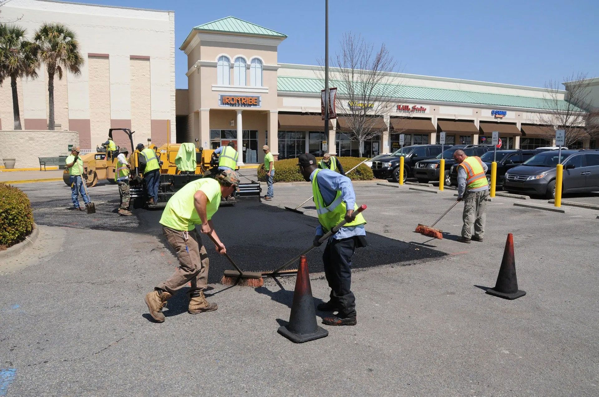 Workers in neon vests repair pavement in a parking lot outside a shopping center.