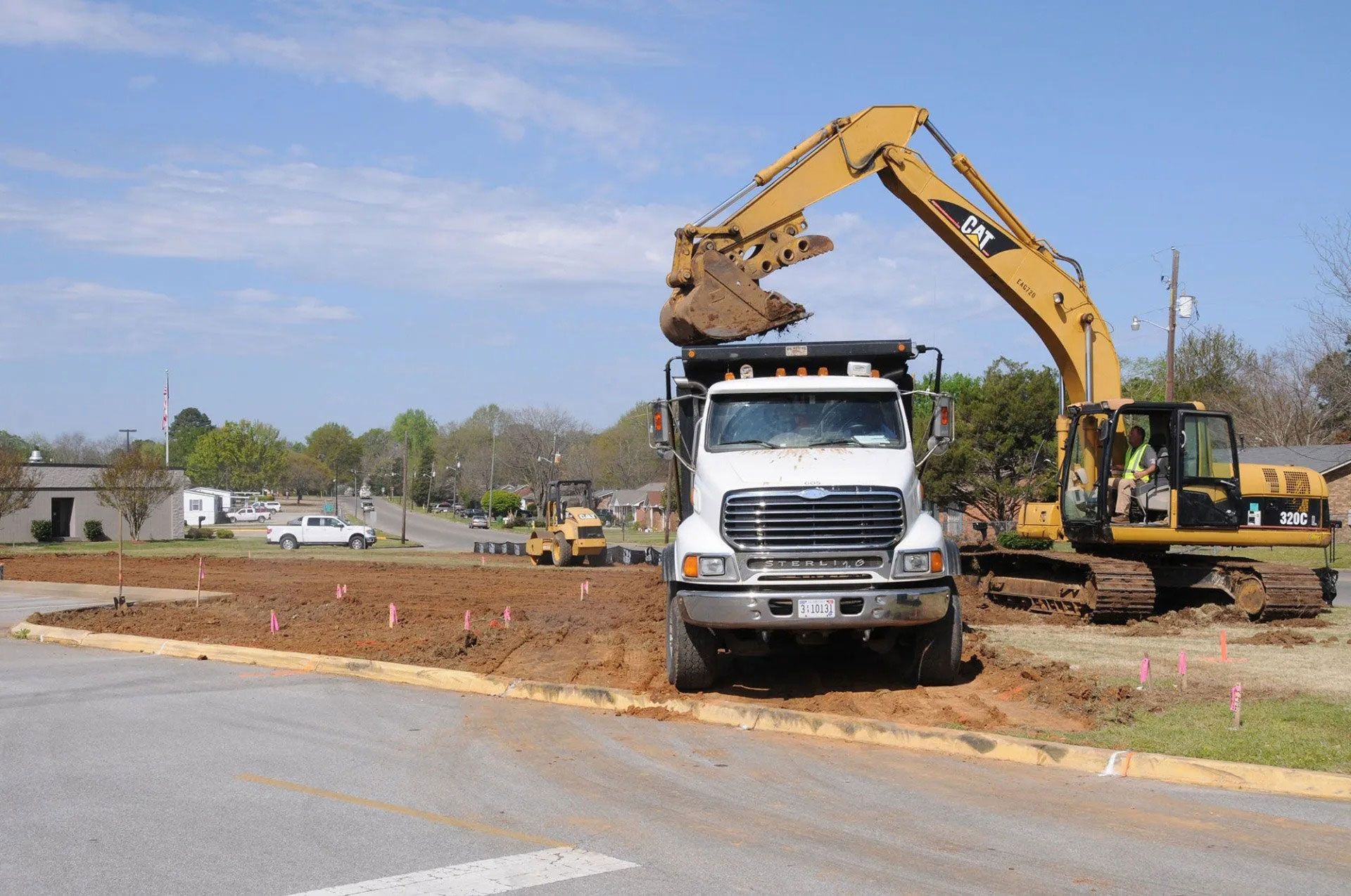 Excavator dumping dirt into a white dump truck at a roadside construction site.