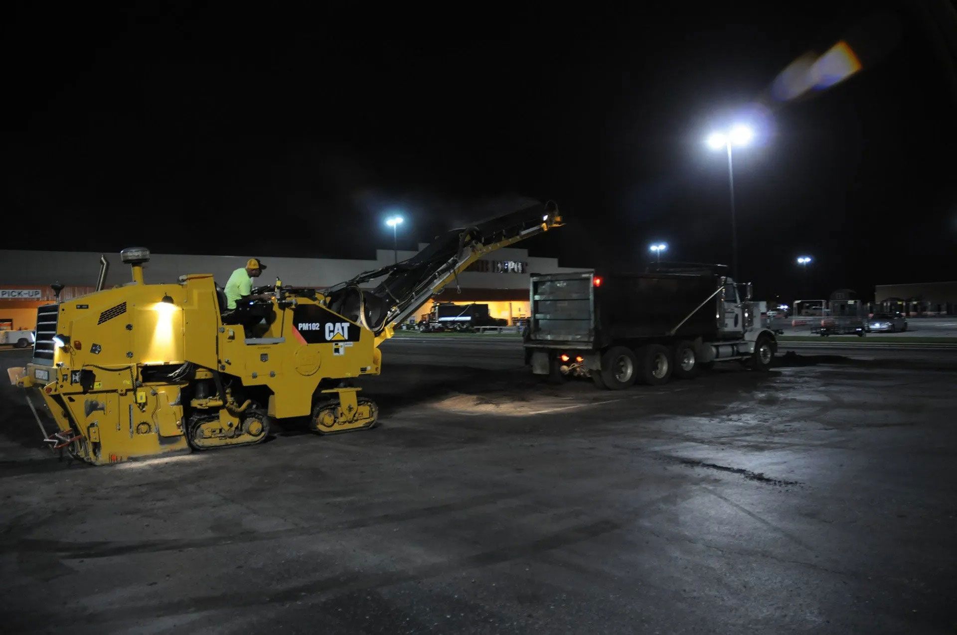 Yellow road milling machine loading asphalt onto a trailer at night under parking lot lights