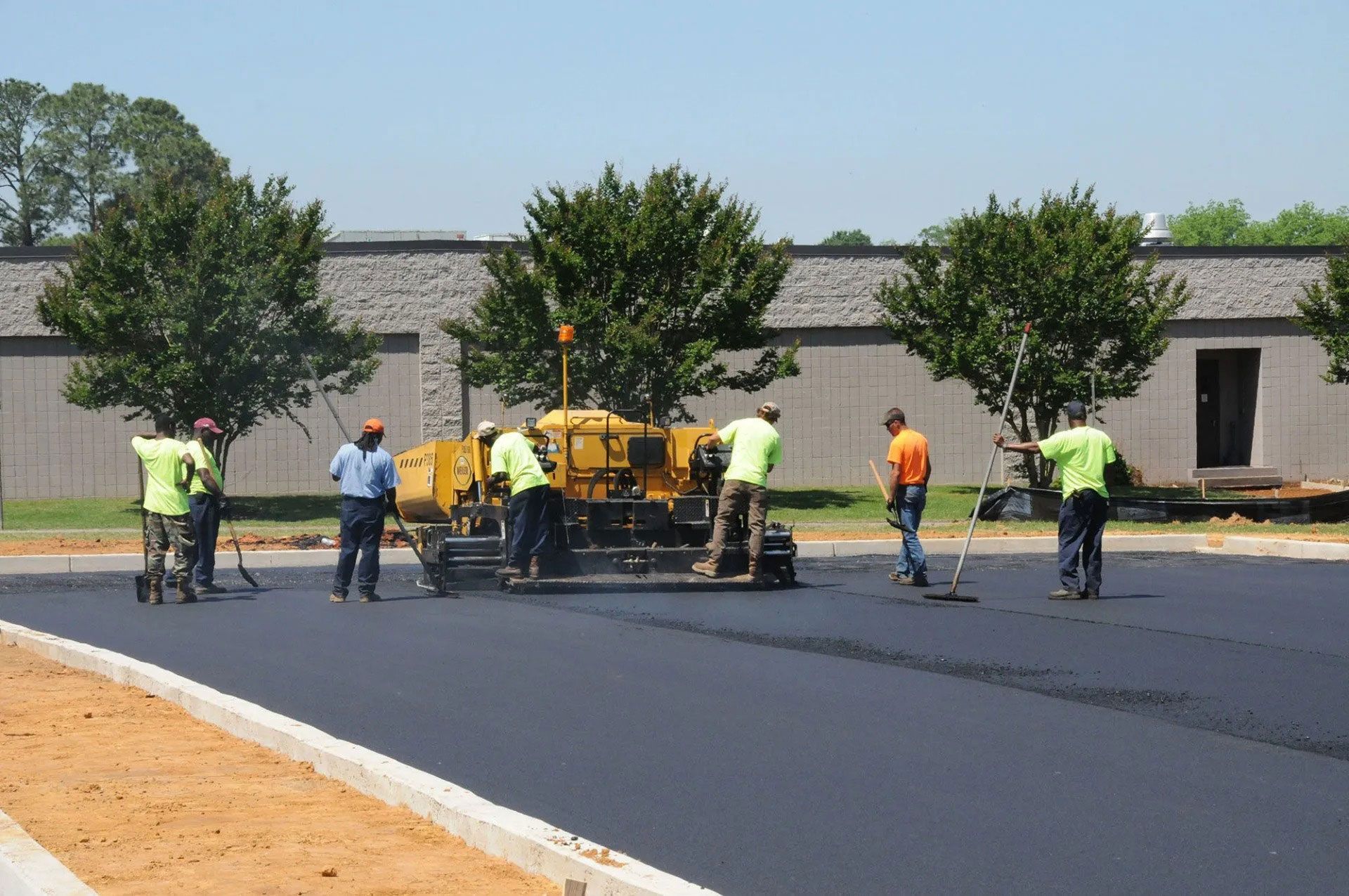 Workers paving a new black road beside a gray wall and trees on a sunny day
