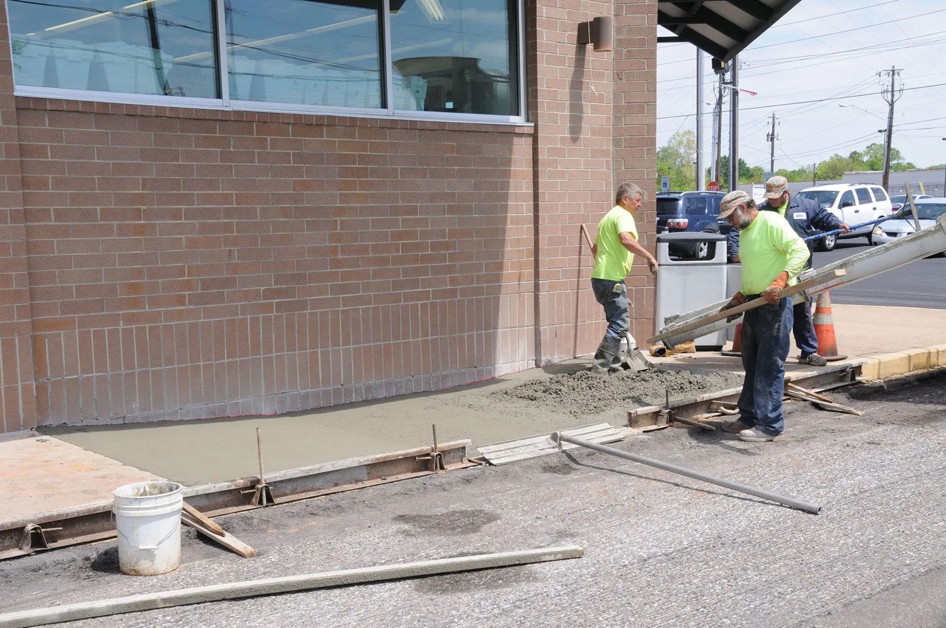 Workers pouring concrete along a building sidewalk beside a parking lot