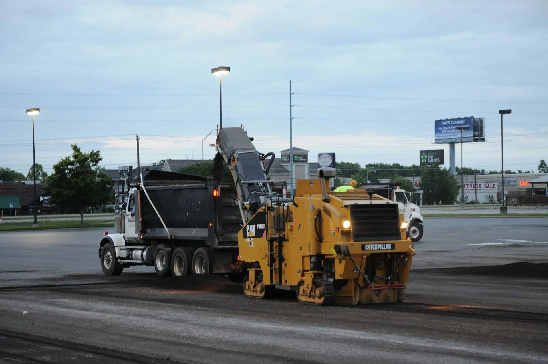 Yellow road milling machine beside a black dump truck on a paved lot at dusk
