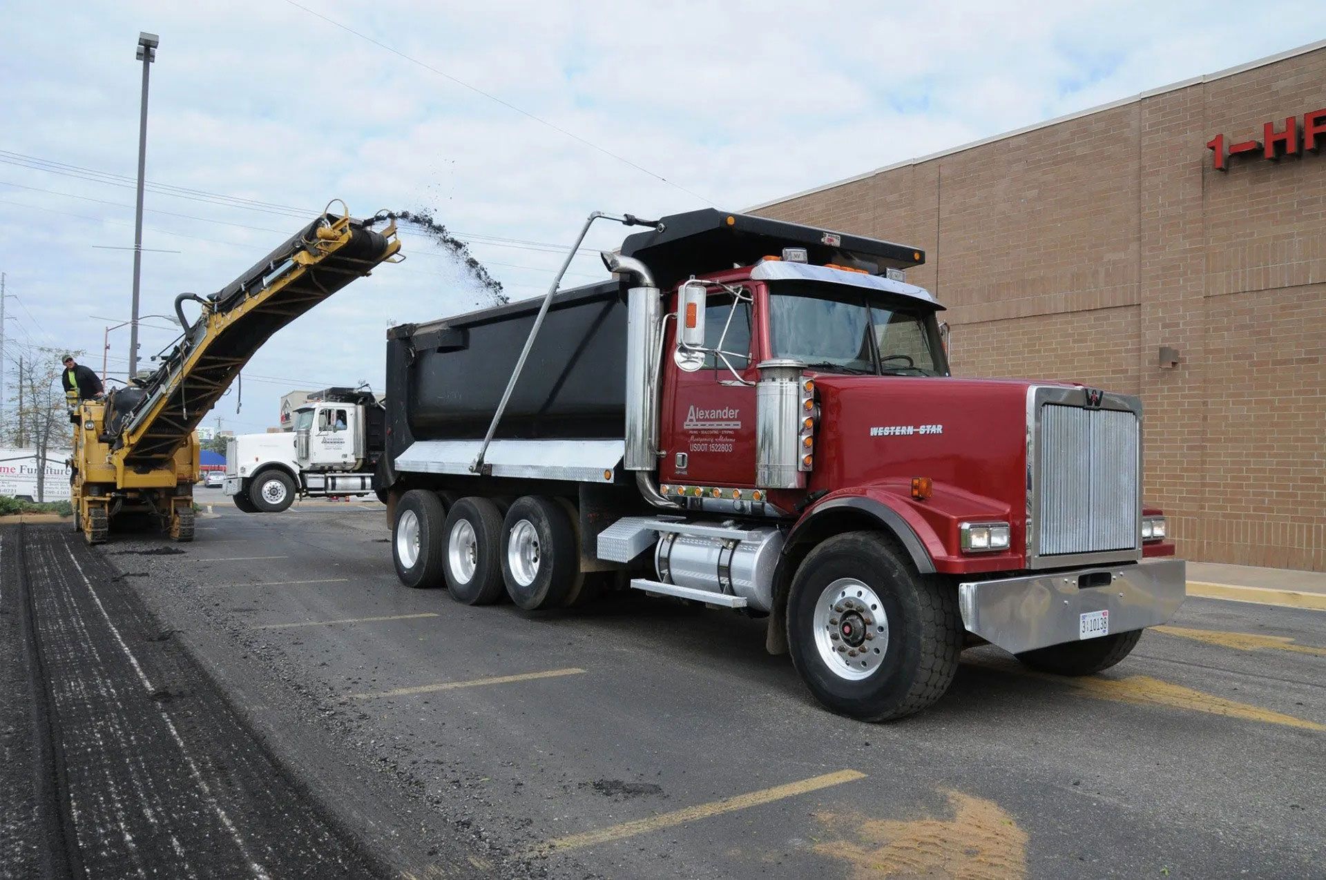 Red dump truck being loaded by a yellow excavator beside a warehouse loading dock.