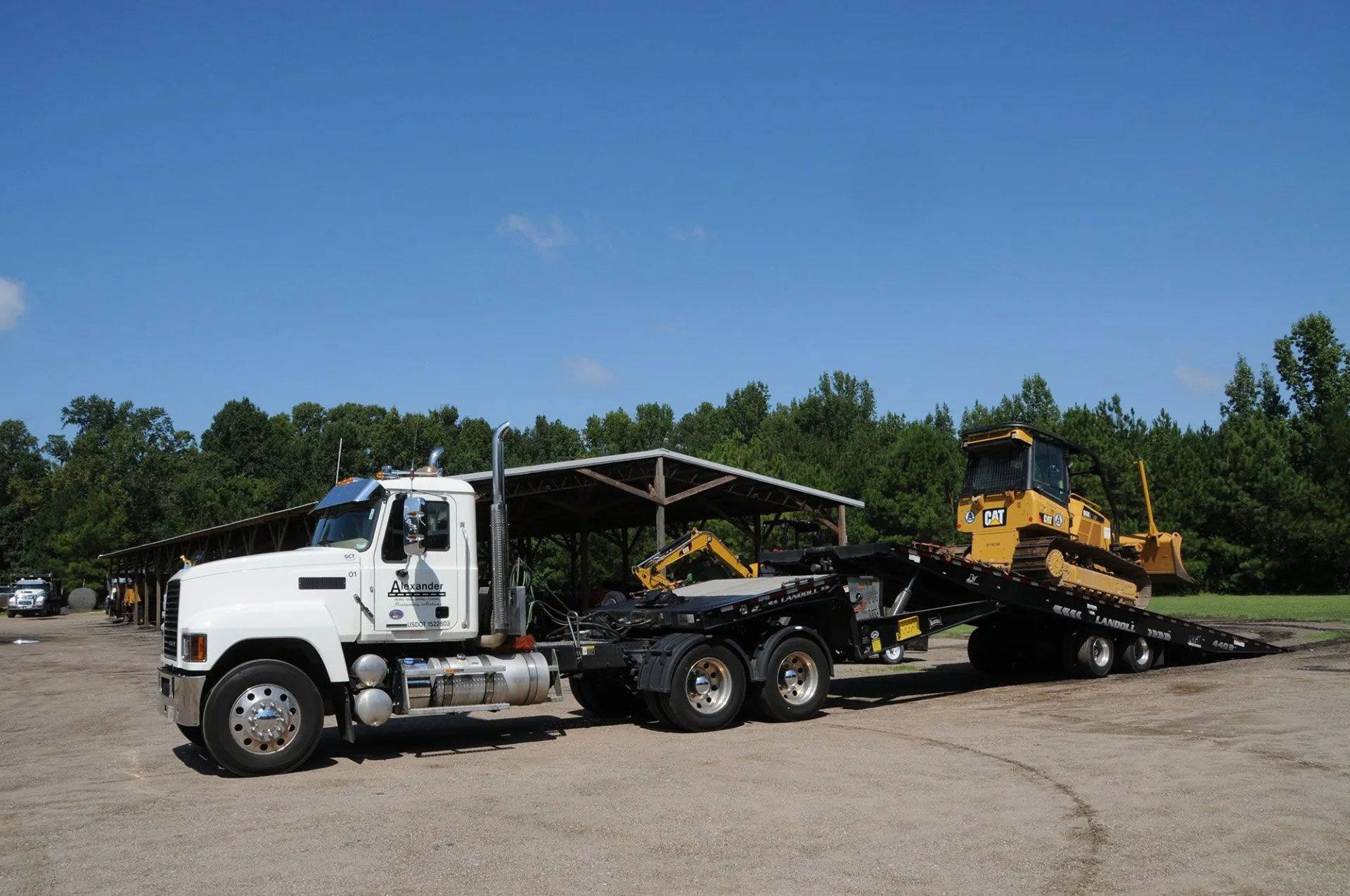 White semi-truck hauling construction equipment on a flatbed trailer in a gravel lot under a blue sky