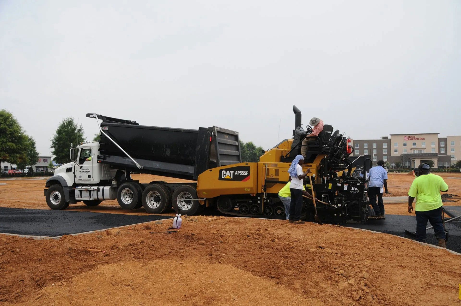 Workers using paving machinery on a road construction site with a dump truck and pile of gravel.