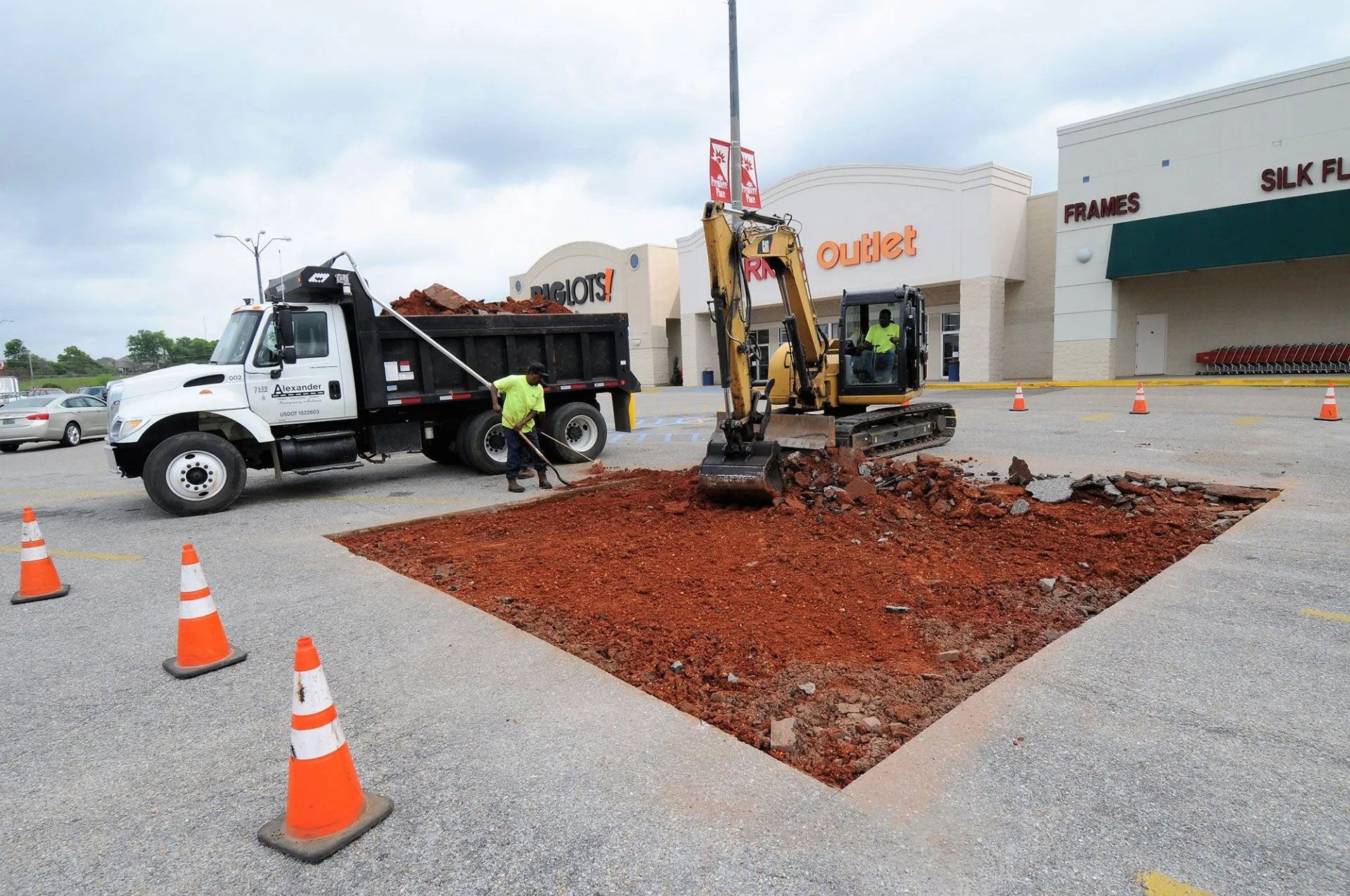 Work truck and excavator repairing a road cutout in a parking lot, with orange cones nearby