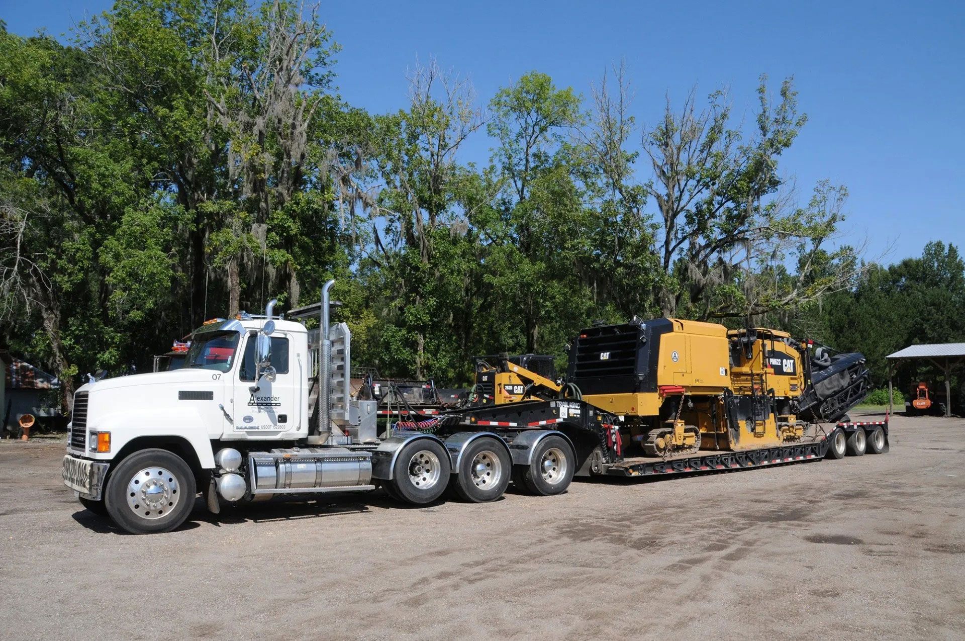 White semi-truck hauling yellow construction equipment on a trailer in a gravel lot