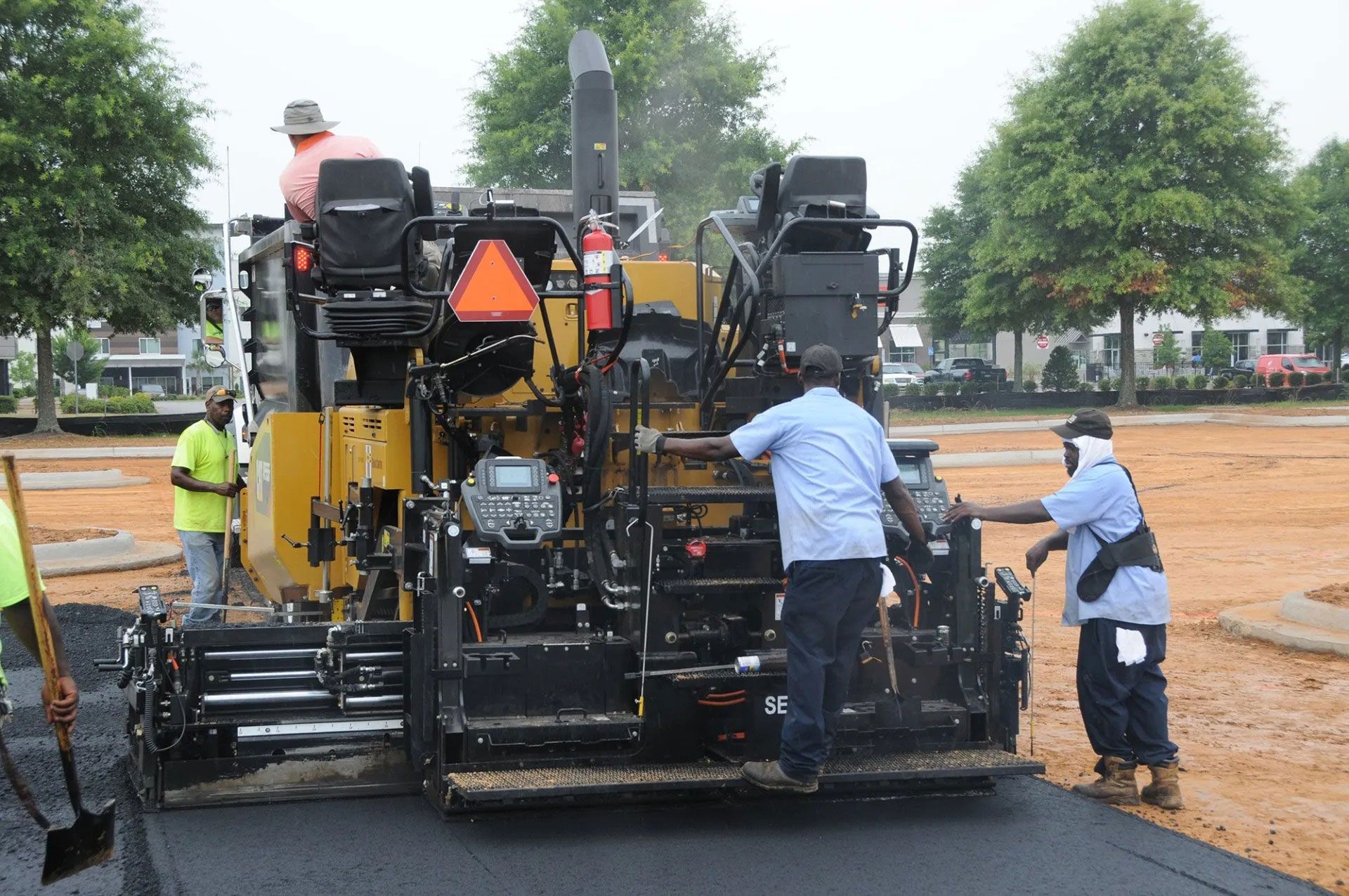 Workers operate a road paver laying fresh asphalt on a wet road surface.