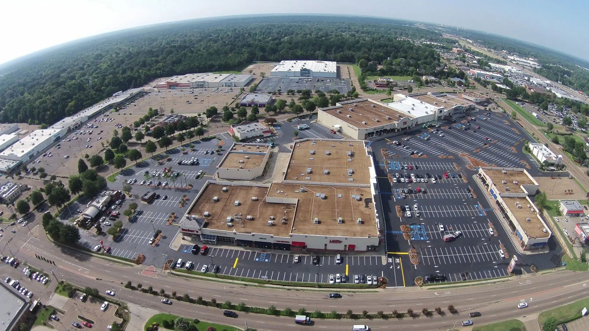 Aerial view of a large shopping center with parking lots, surrounding roads, and trees in the background