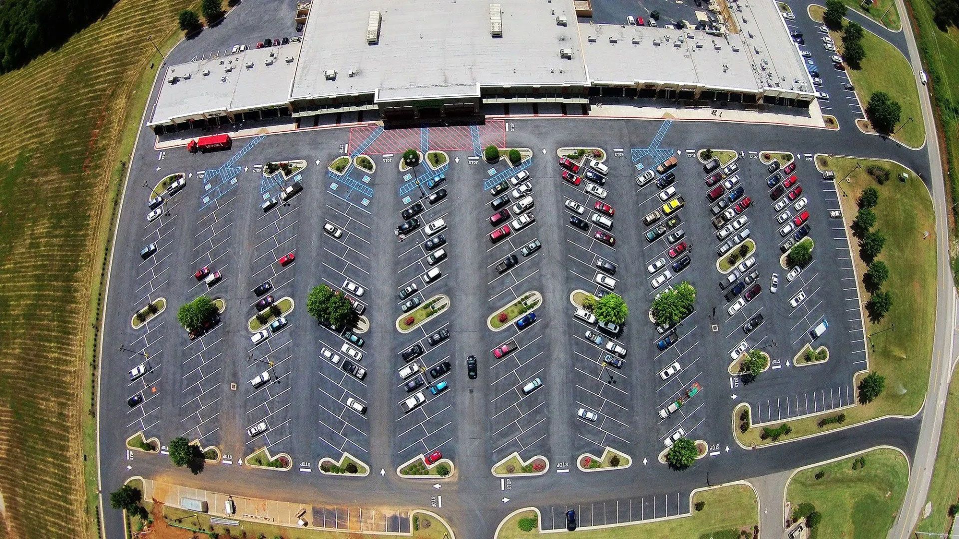 Aerial view of a crowded parking lot beside a large building, with rows of parked cars and surrounding greenery.