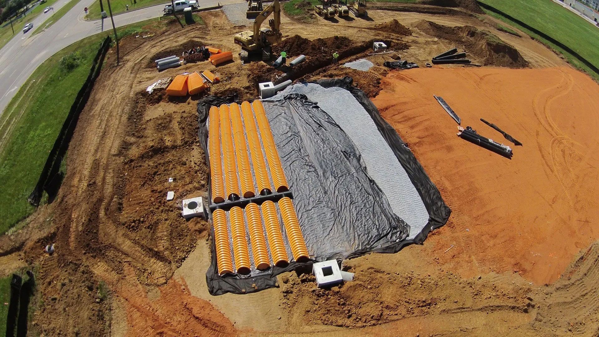 Aerial view of a construction site with orange soil, gravel beds, and equipment near a road and fields