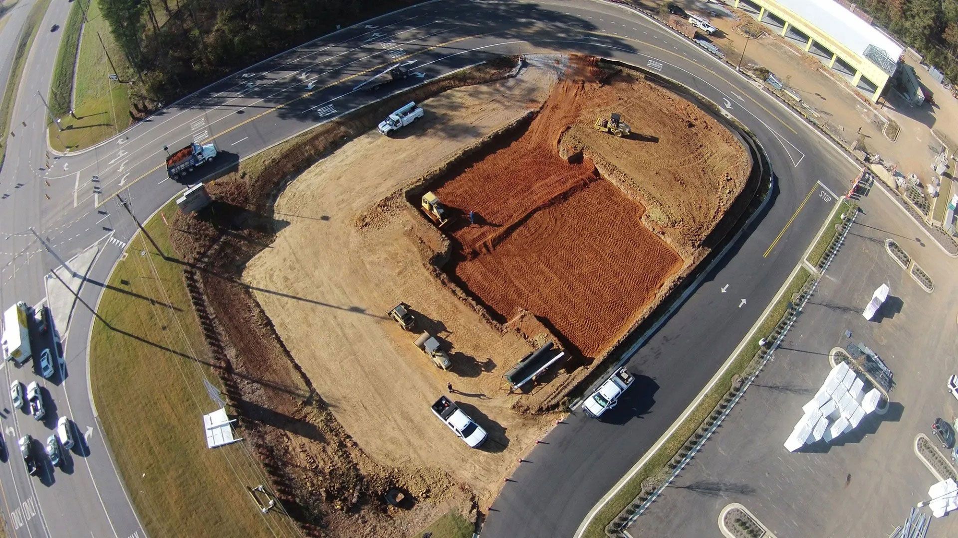 Aerial view of a construction site with dirt piles, parked vehicles, and surrounding roads.