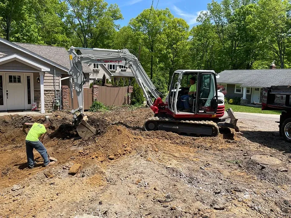 Construction site: Excavator digging in front yard of a house while a worker shovels soil.