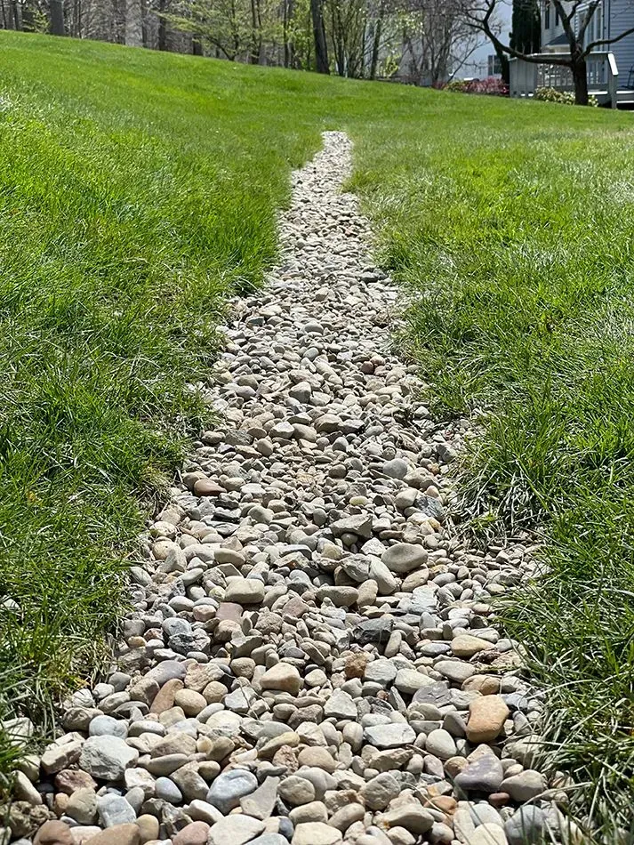 A gravel-filled drainage channel cuts through a grassy lawn.