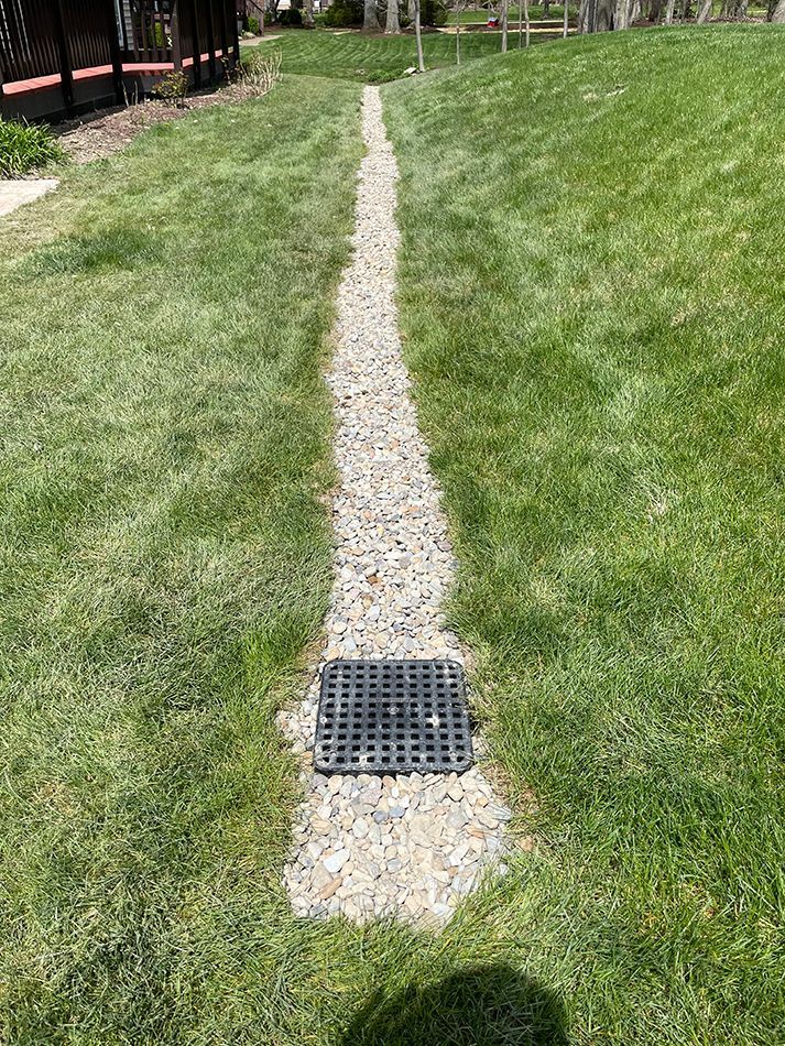 Gravel-lined drainage channel in a grassy yard, leading to a square grate.