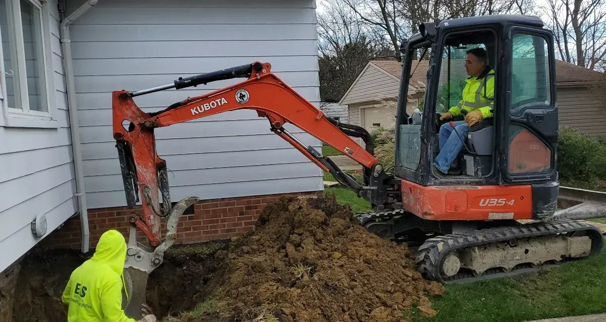 Man operating mini excavator digging near a house, another worker in yellow vest watches.