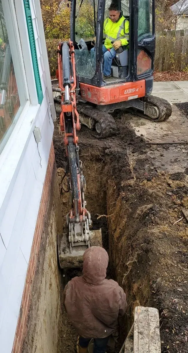 Mini excavator and worker in a trench alongside a building, digging near a window.