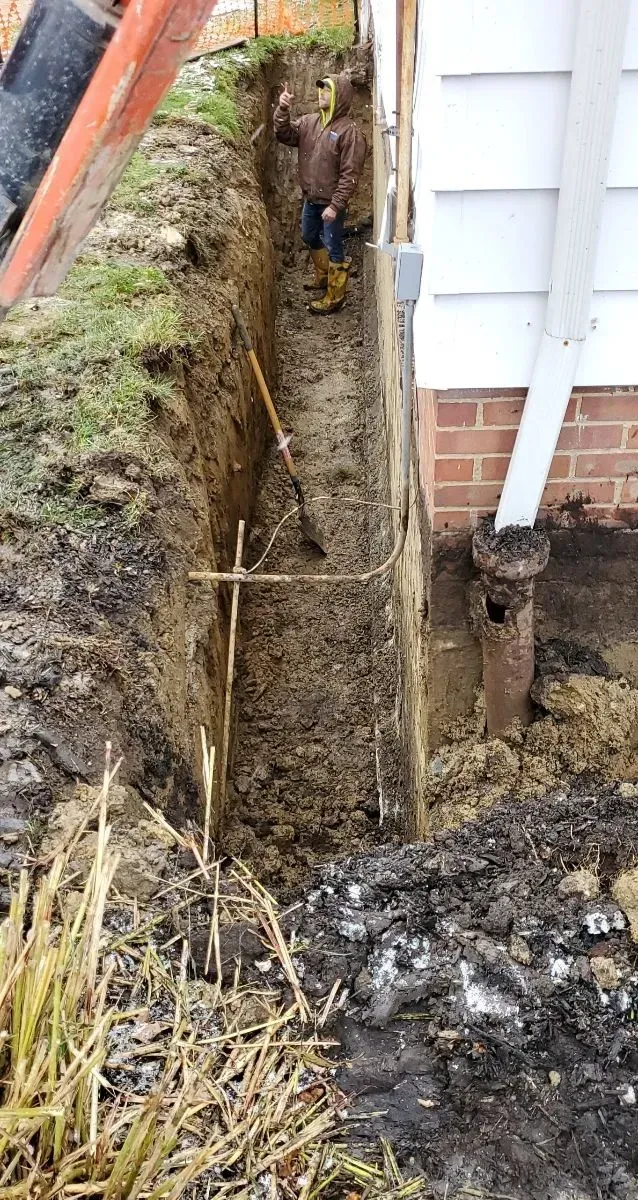Man in trench next to house digging with shovel; brown soil, white siding, downspout.