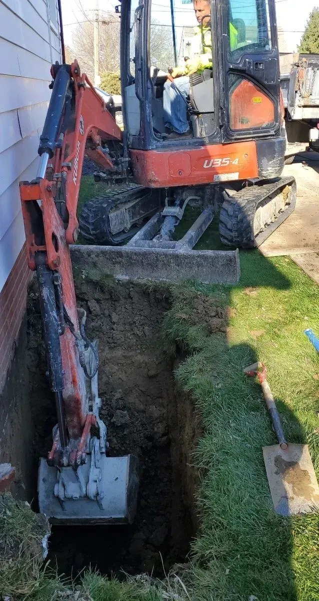 Mini excavator digging a trench along a building. Orange machine, dirt, and green grass.