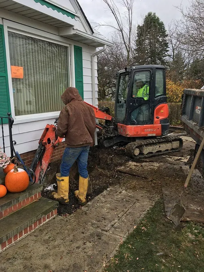 Person operating excavator digging near a house, orange pumpkins on steps.